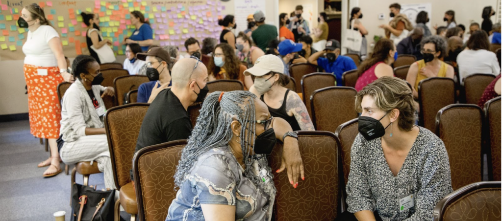 People of various ages and races wearing black N95 masks and seated in closely placed upholstered auditorium chairs.